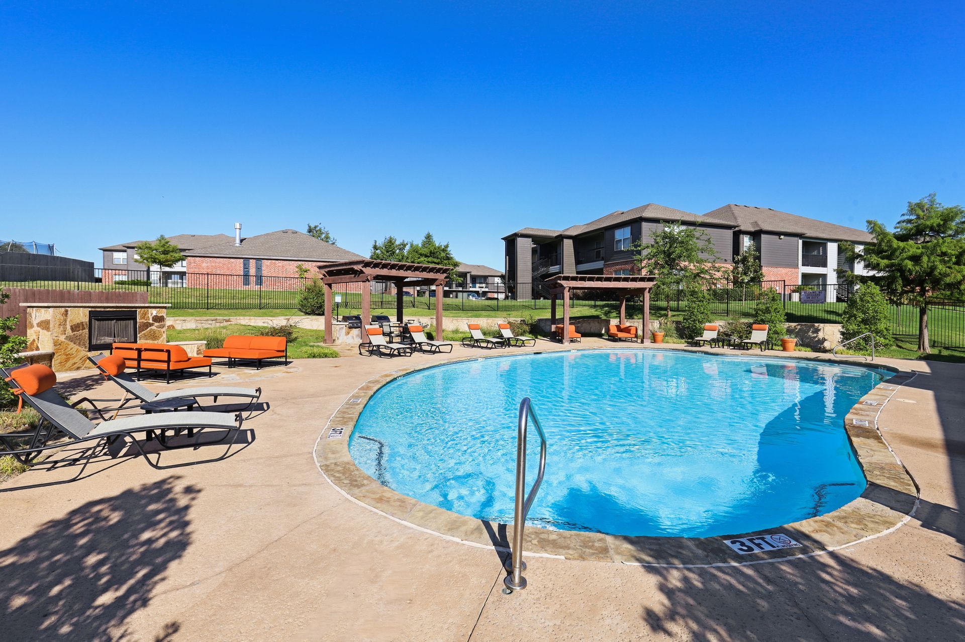 Pool area with lounge chairs, pergolas, and buildings under a blue sky.