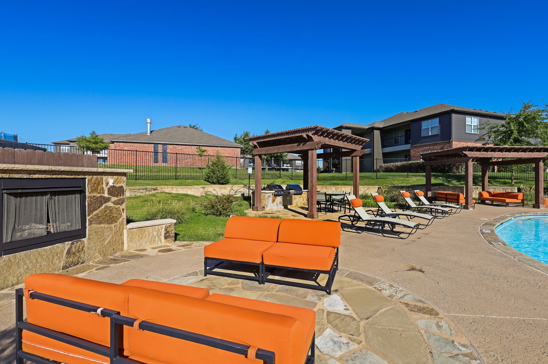 Outdoor seating area with orange couches, pool, and buildings under a blue sky.