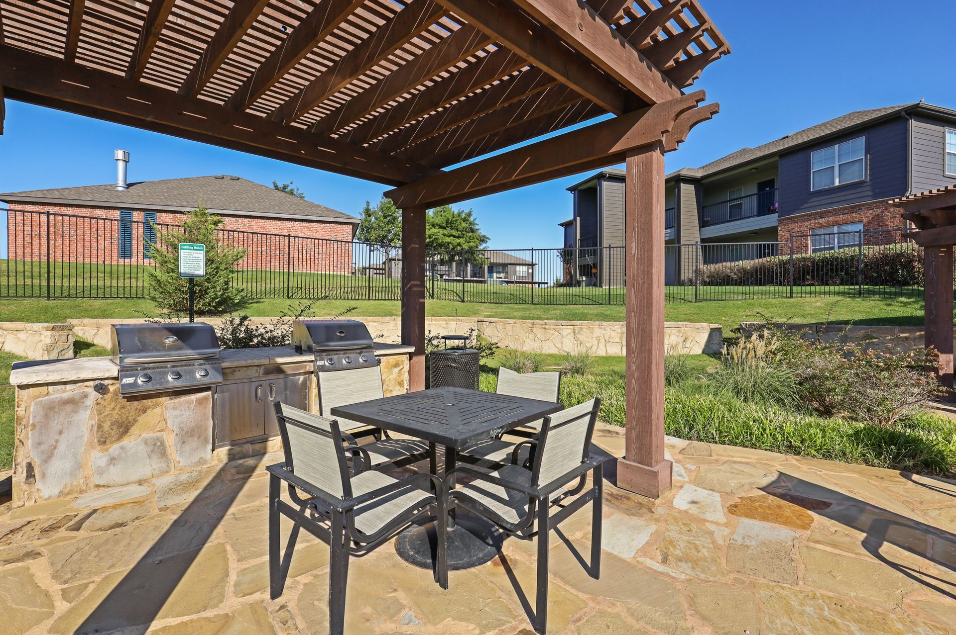 Outdoor grilling area with a pergola, stone patio, grills, table, chairs, and surrounding buildings.