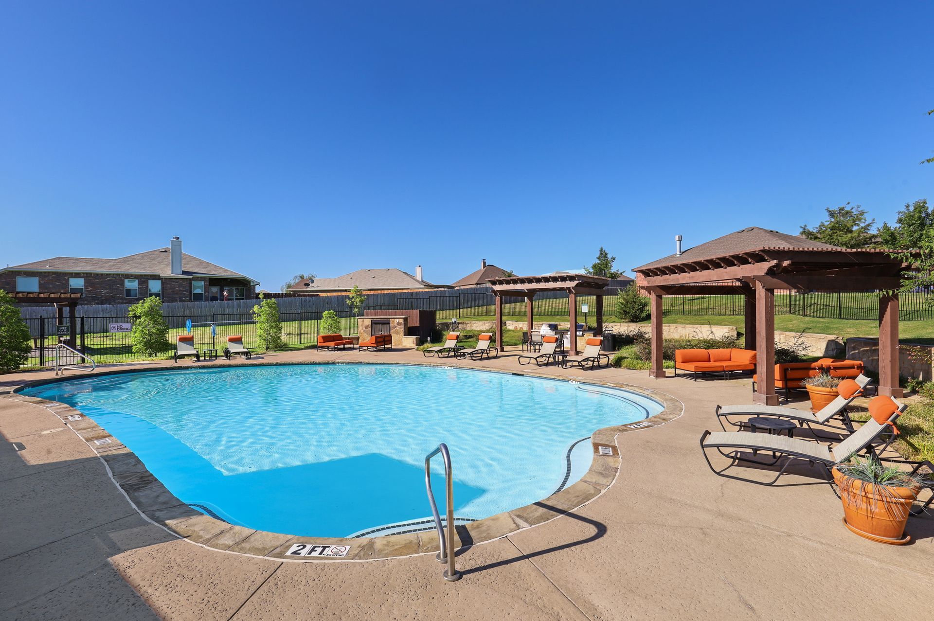 Swimming pool with lounge chairs, gazebos, and houses under a clear blue sky.