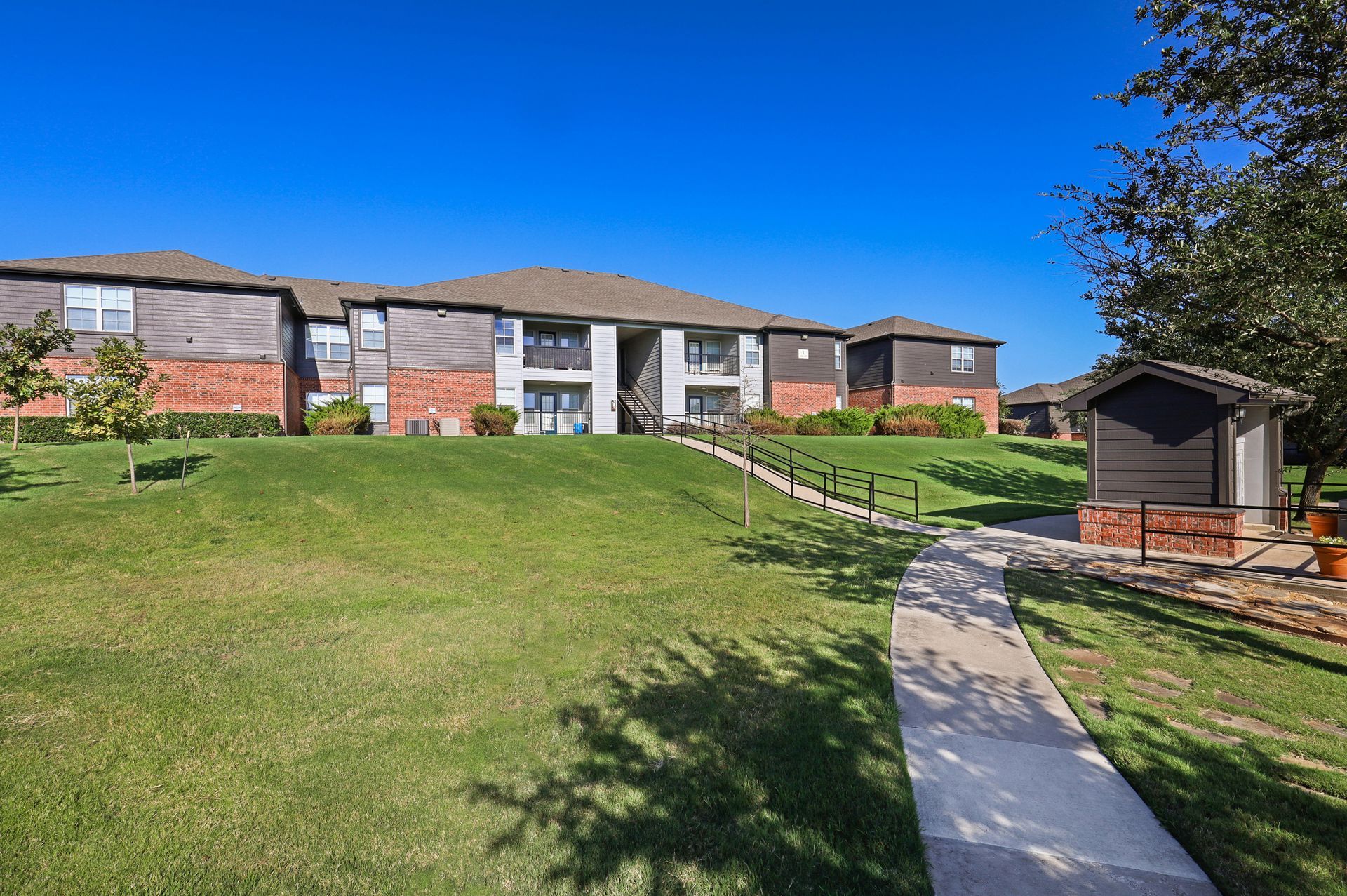 Apartment buildings with red brick, brown siding, and a grassy lawn, pathway, and blue sky.