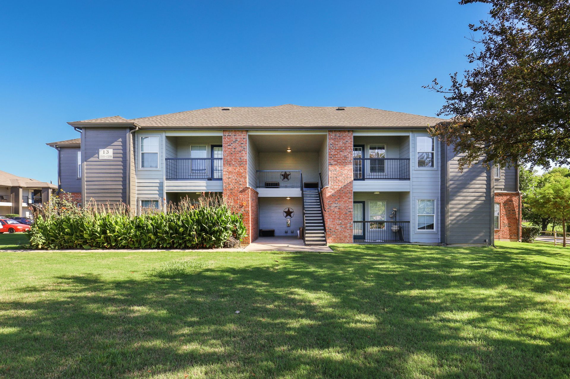 Two-story apartment building with brick accents and balconies on a grassy lawn under a blue sky.