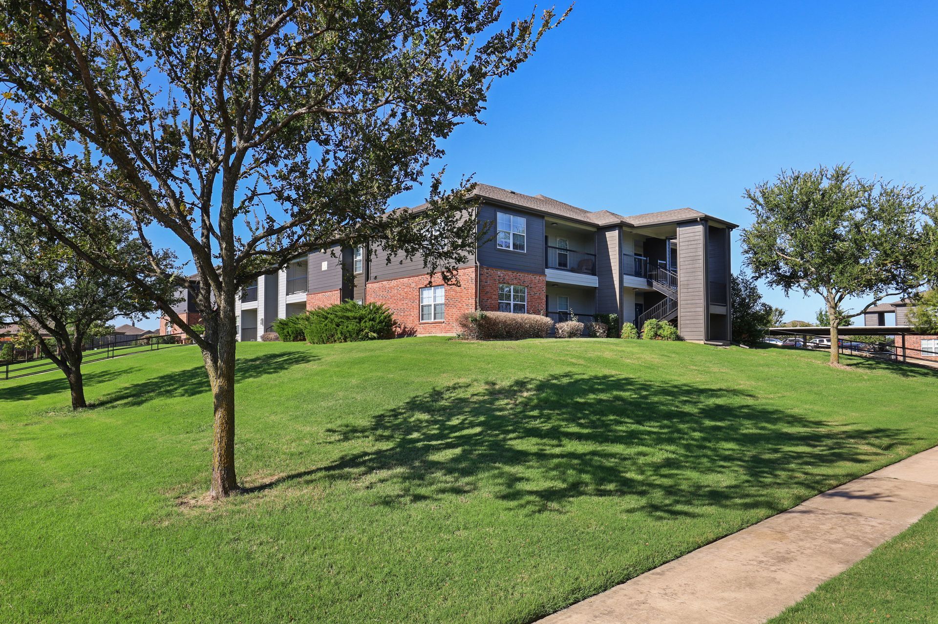 Apartment building on a grassy hill, surrounded by trees under a blue sky. A sidewalk is in the foreground.