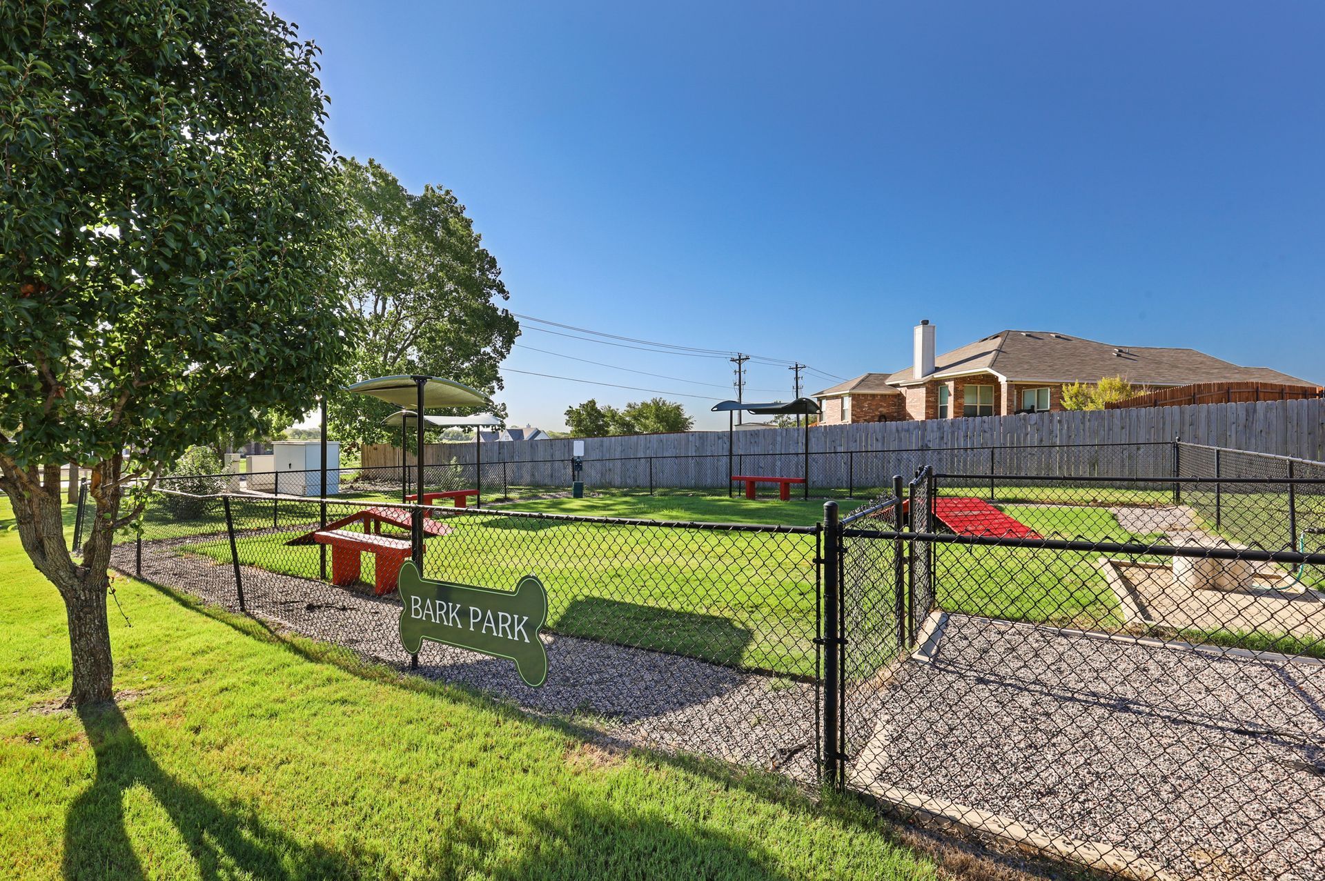 Fenced dog park with green grass, picnic tables, and agility equipment under a blue sky.