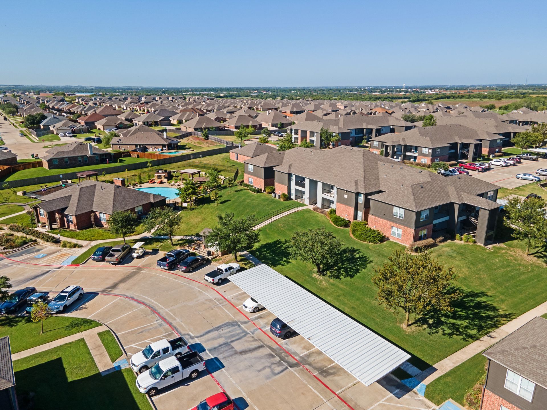 Aerial view of apartment complex with pool, trees, and carports in suburban setting.