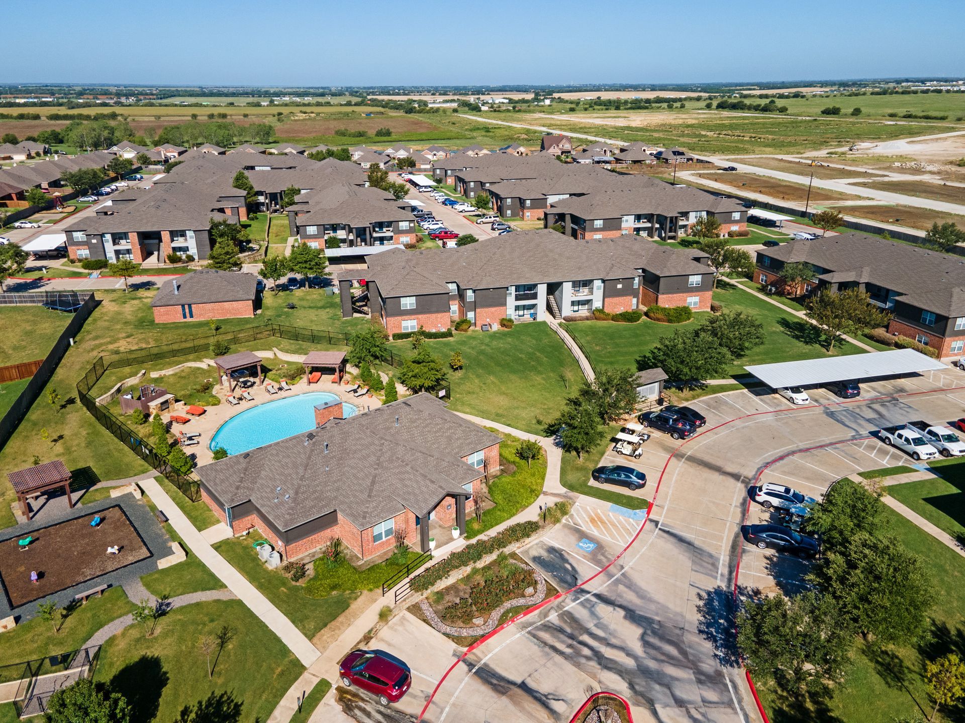 Aerial view of an apartment complex with brown roofs, a pool, and green lawns on a sunny day.