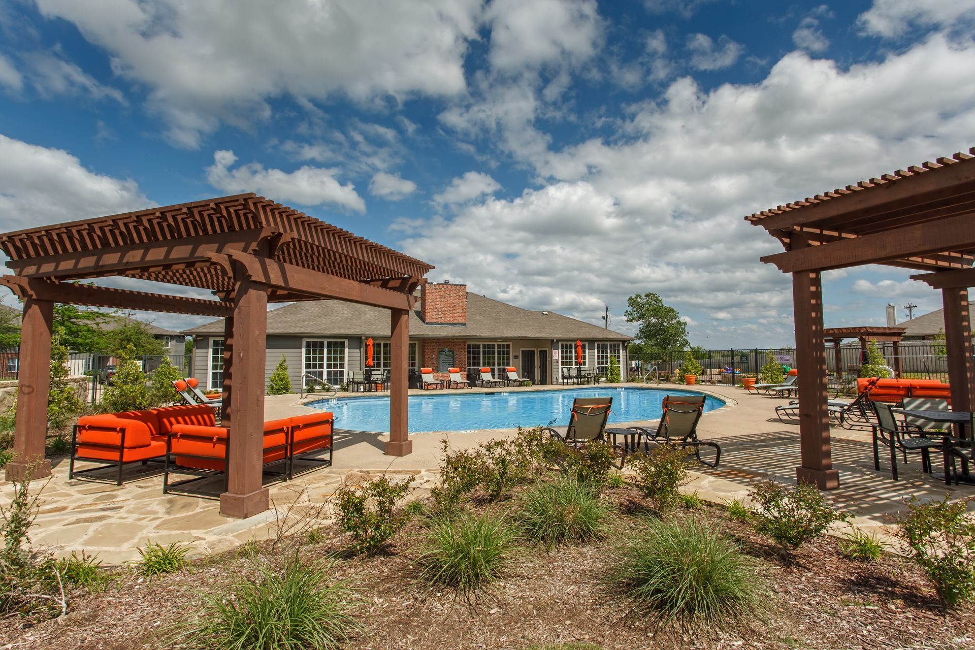 Pool area with brown pergolas, lounge chairs, and blue pool under a cloudy sky.