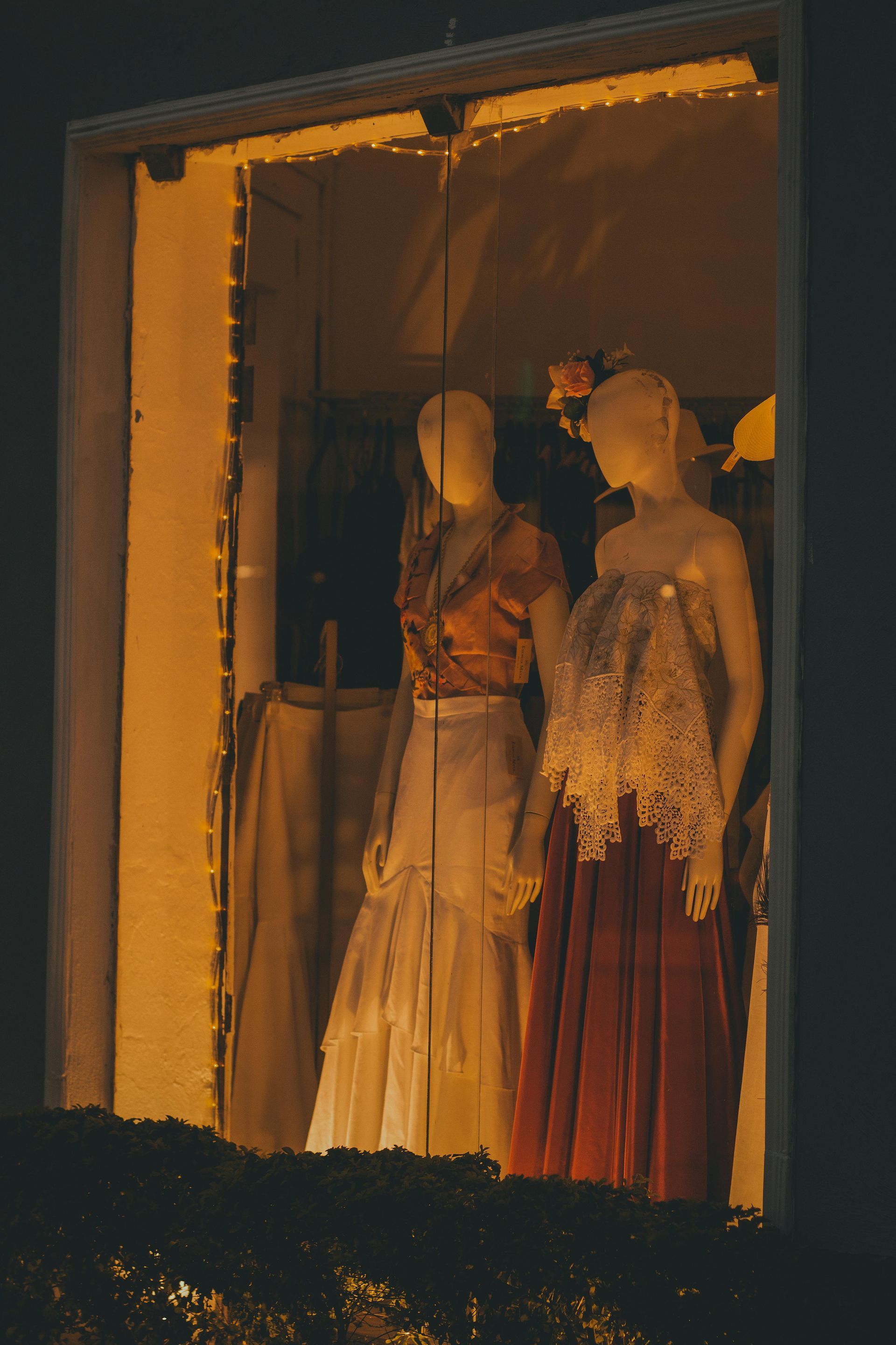 Two mannequins in a well-lit store window display; one in a red skirt and lace top, the other in an orange shirt and white skirt.