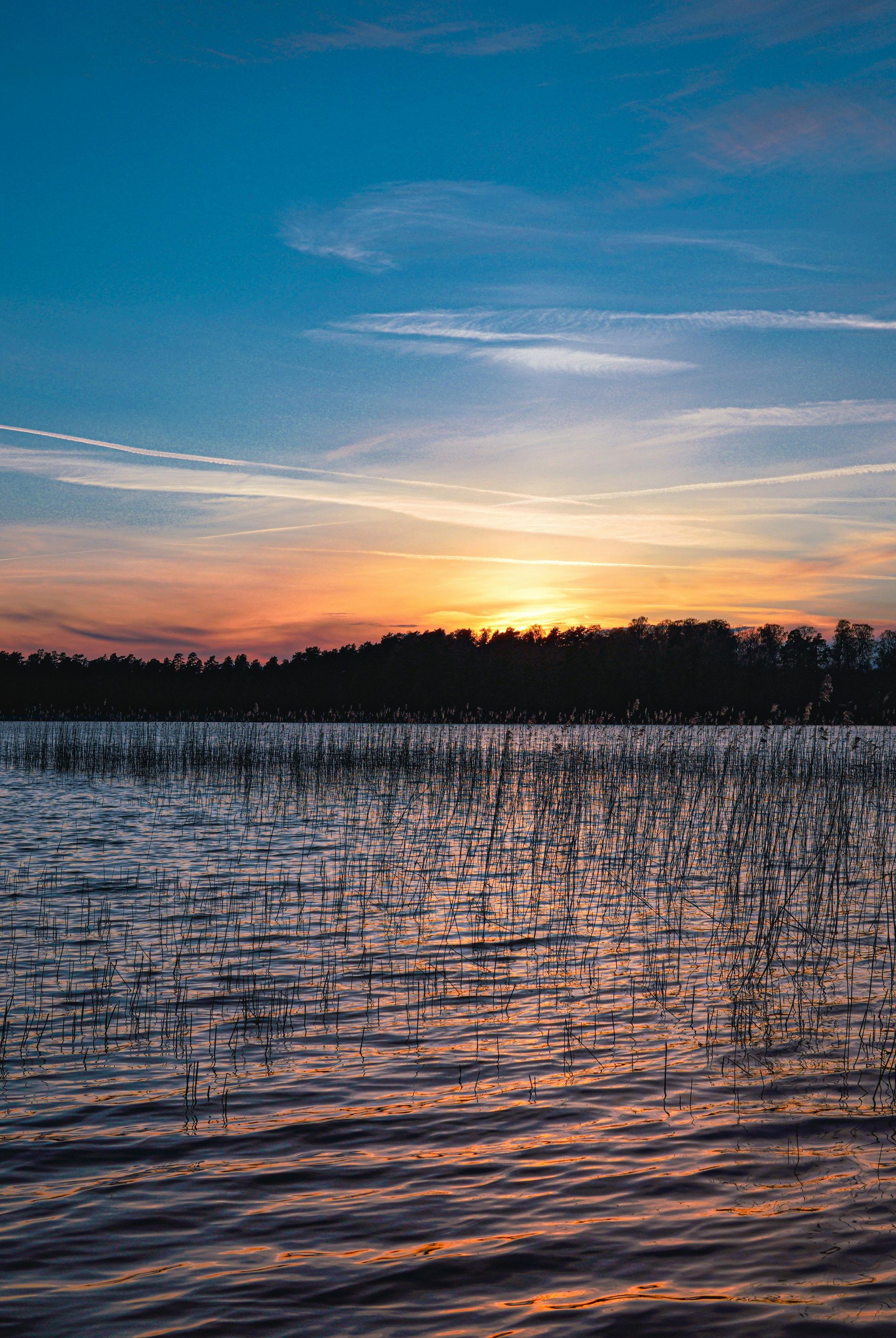 Sunset over a lake; orange and yellow hues above dark tree line, water with vertical reflections under blue sky.