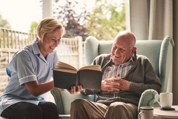 Caregiver and senior man laughing while reading a book in a sunlit room, teal chair.