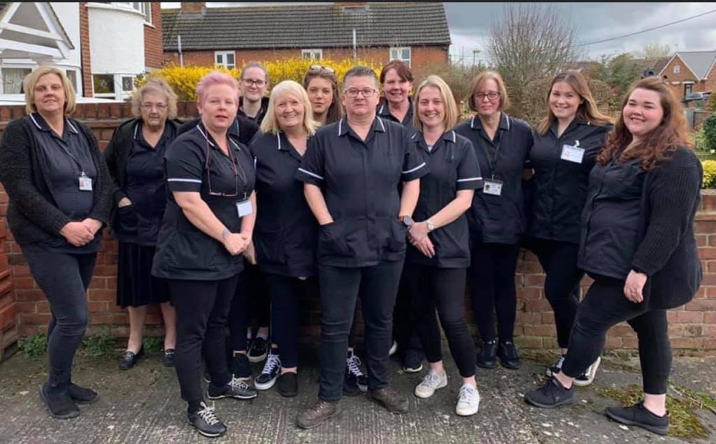 Group of people in black uniforms posing outdoors near a brick wall and a building with a cloudy sky.