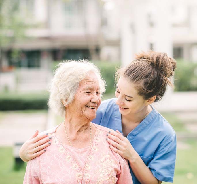 Young woman in blue scrubs smiles at older woman with hands on her shoulders outdoors.