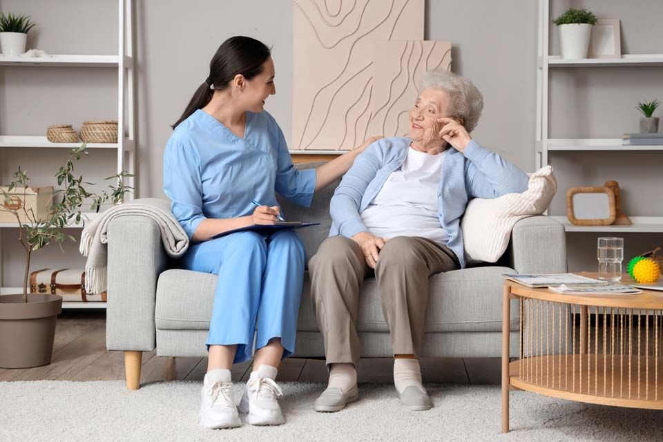 A caregiver in blue scrubs talks to a seated senior; both on a couch. Living room setting, shelves in background.