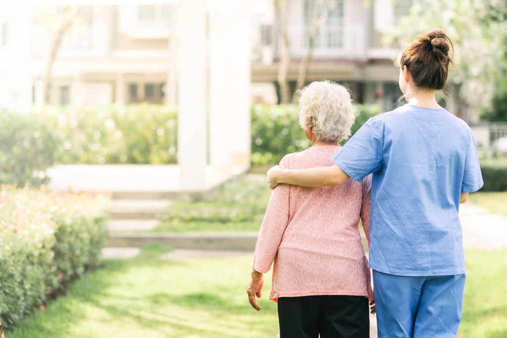 Caregiver in blue uniform assists elderly person walking outdoors.
