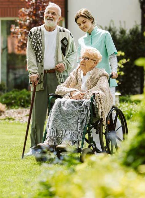 Man and woman in a wheelchair outside, with a caregiver. The man holds a cane, all looking towards something.
