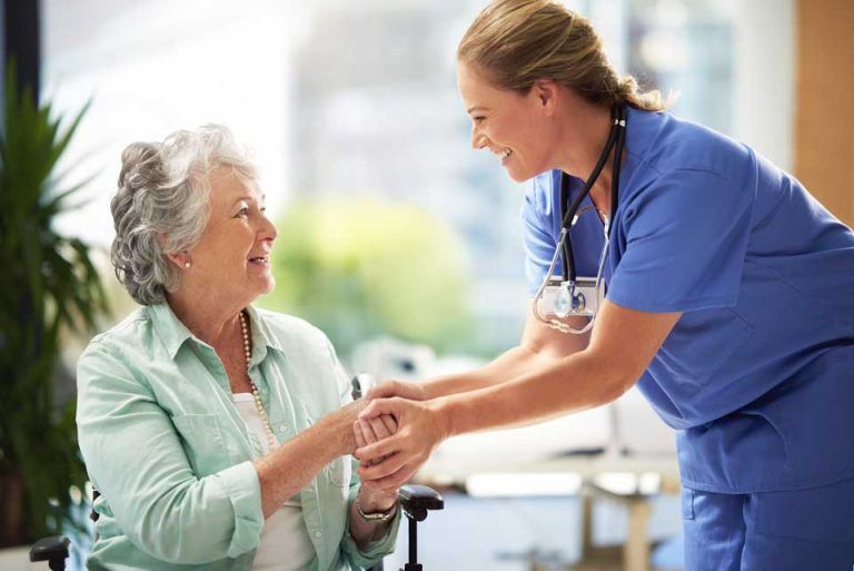 Nurse assisting a smiling older person in a wheelchair, holding hands. Indoors, natural light.