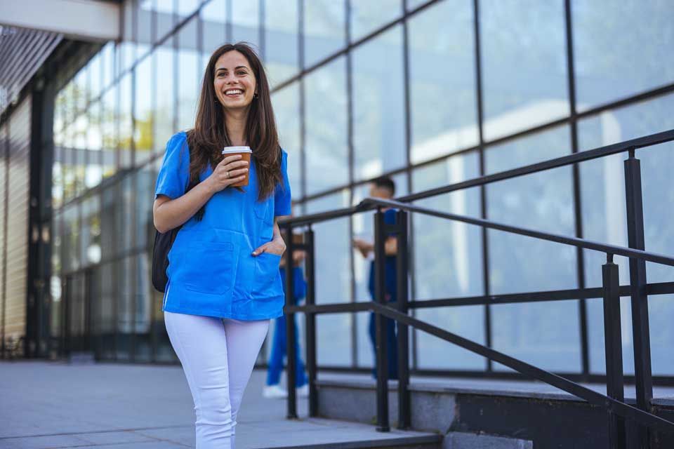 Woman in blue scrubs and white pants holding coffee, smiling in front of a building with glass windows and a metal railing.