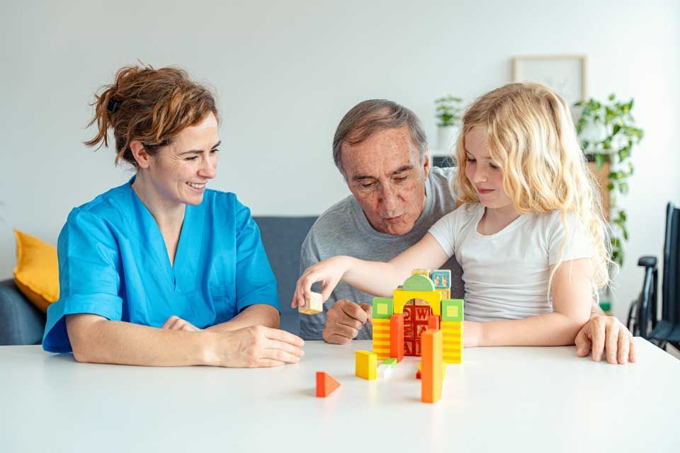A young child, older adult, and healthcare worker play with colorful building blocks at a table.