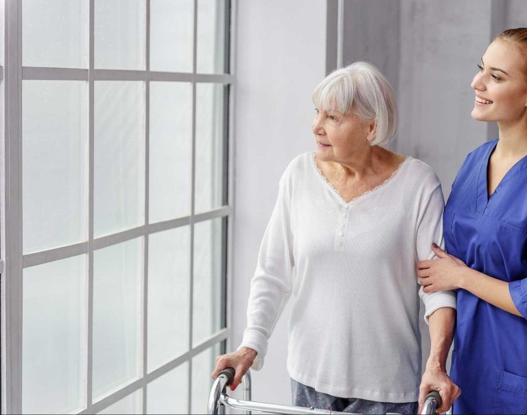 Elderly woman with walker looking out window, assisted by caregiver in blue scrubs.