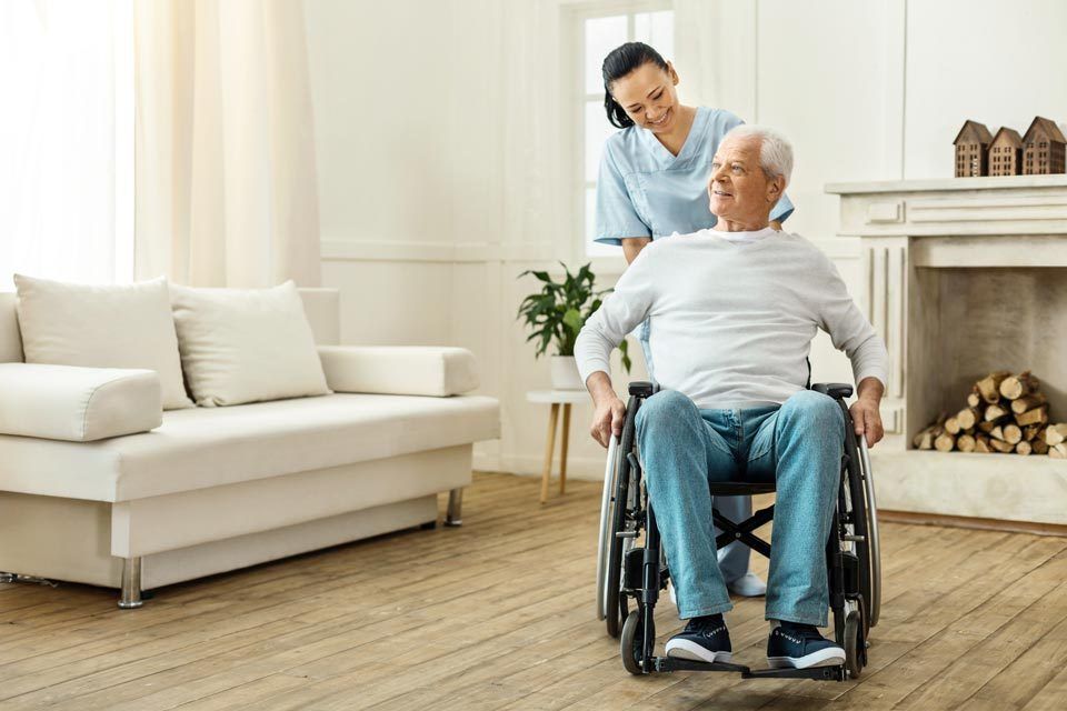 Caregiver pushing a man in a wheelchair indoors near a couch and fireplace.