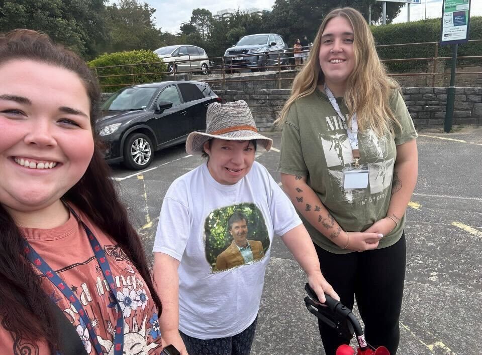 Three people smiling outdoors near parked cars; one person uses a mobility aid.
