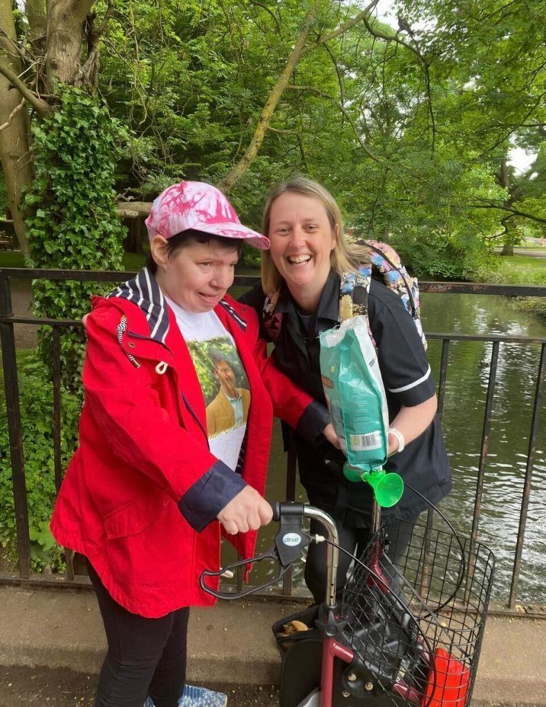 Two people smiling by water, one with a walker, holding food bag, other wearing a pink hat and red jacket.