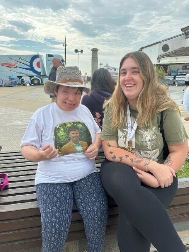 Two people smiling outside. One wears a hat and shirt with a photo. Other in black leggings, olive shirt.