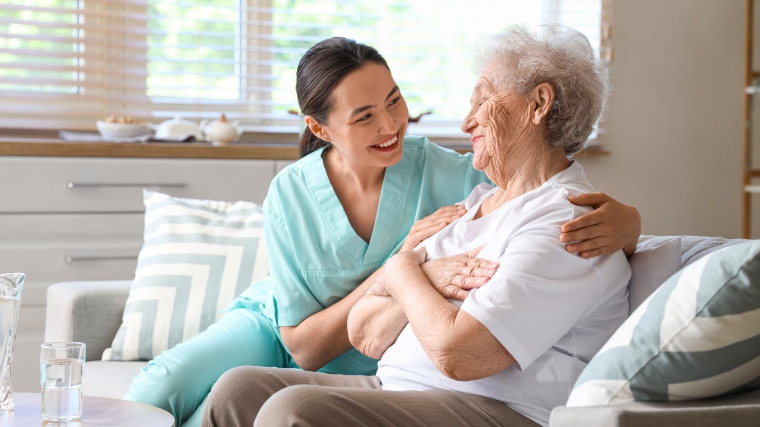 Caregiver in teal scrubs comforts an elderly person on a couch in a home setting.