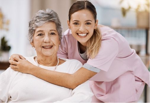 Young caregiver with arm around smiling older person. Both look at the camera. Soft lighting.