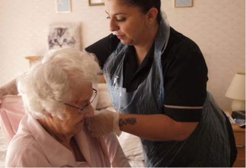 Caregiver assisting an older person, possibly cleaning the face. Indoors, bedroom setting, light skin tones.