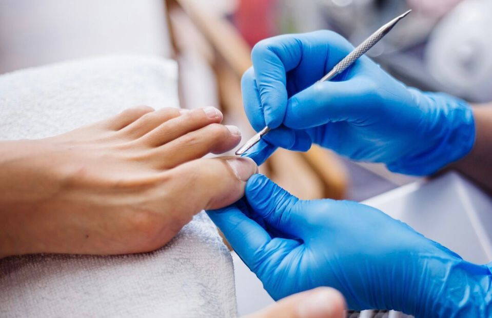 Person's foot being worked on with a nail tool by gloved hands, in a salon setting.