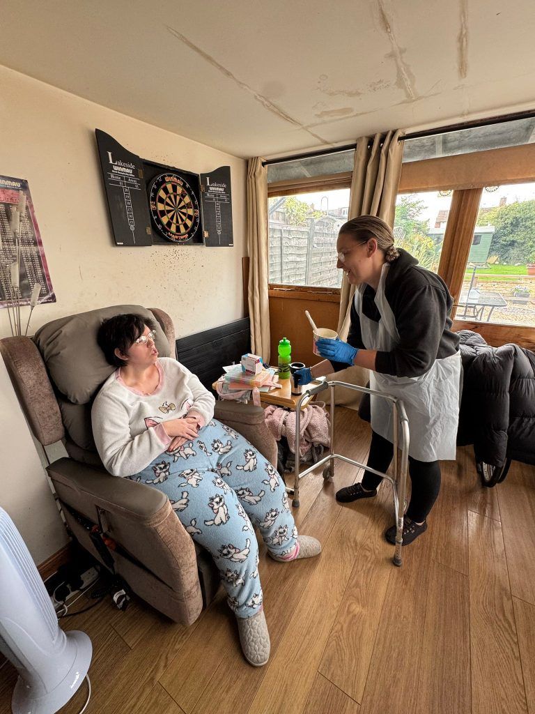 Person in pajamas sits in recliner while another offers food on a tray. Indoor setting with dartboard.