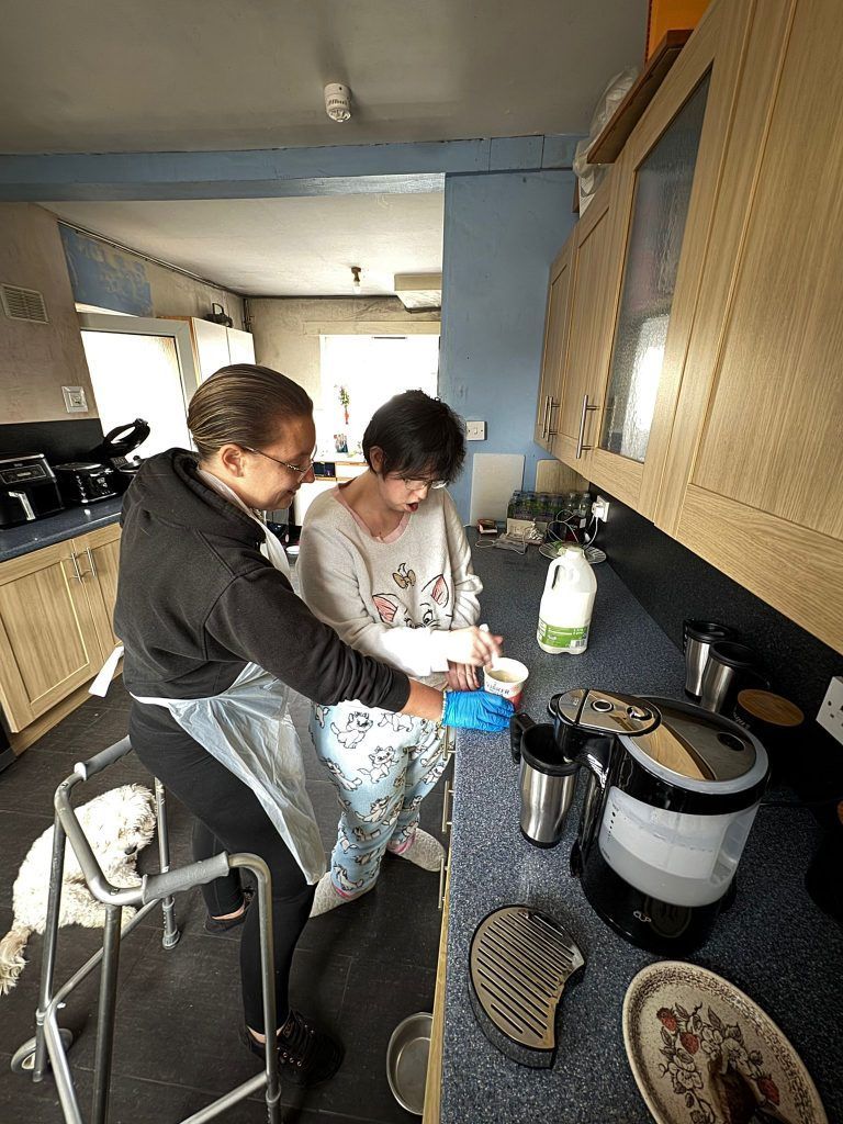 Woman helping another person stir in kitchen, with milk carton, mugs, and a walker.