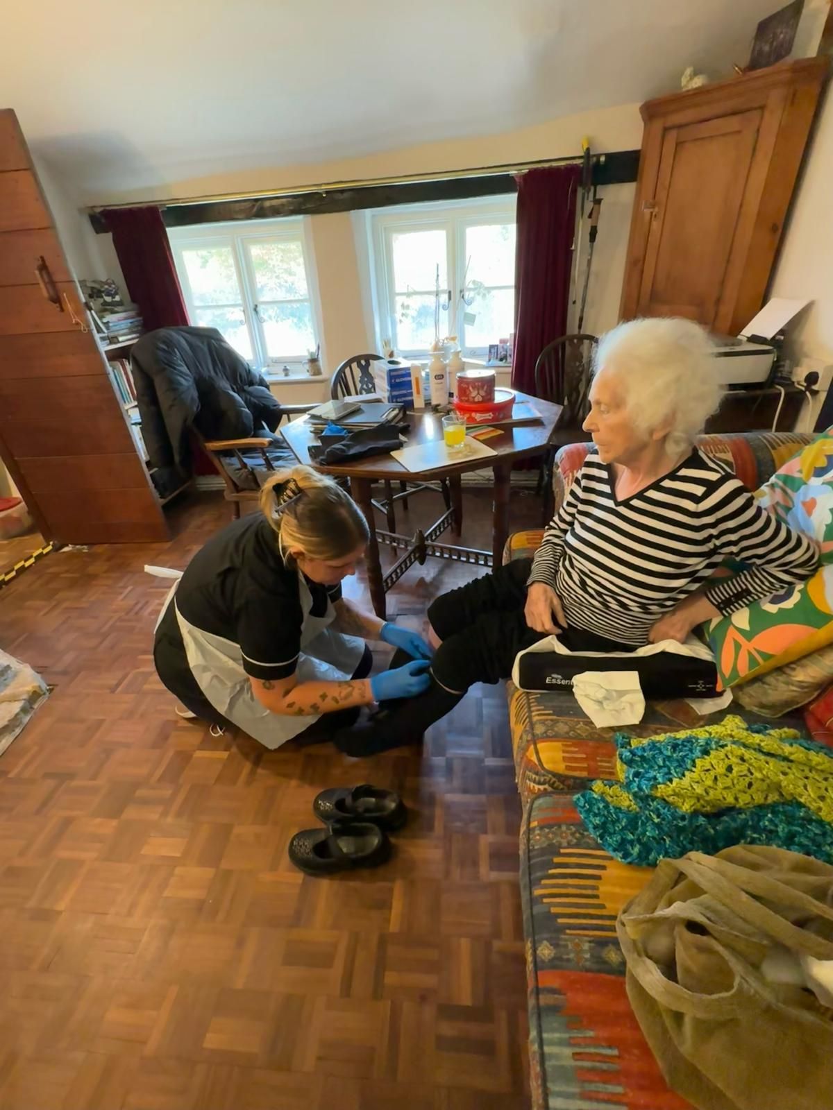 Caregiver tending to an elderly person's foot indoors, wearing black uniform and gloves.
