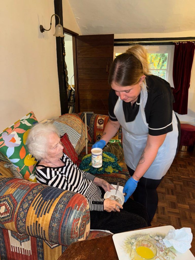 Woman helping elderly person in a home setting; serving a beverage, wearing gloves and apron.