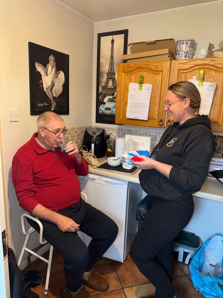 Man drinking water sits in a kitchen; woman leans on counter, holding something, smiling.