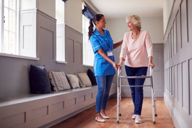A caregiver assists a person with a walker in a hallway. The caregiver has their hand on the other's arm.