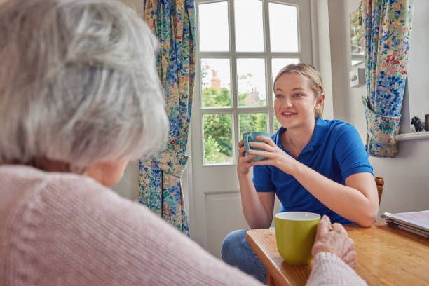 Woman in blue shirt talks to elderly person over a table, holding a mug indoors by a window.