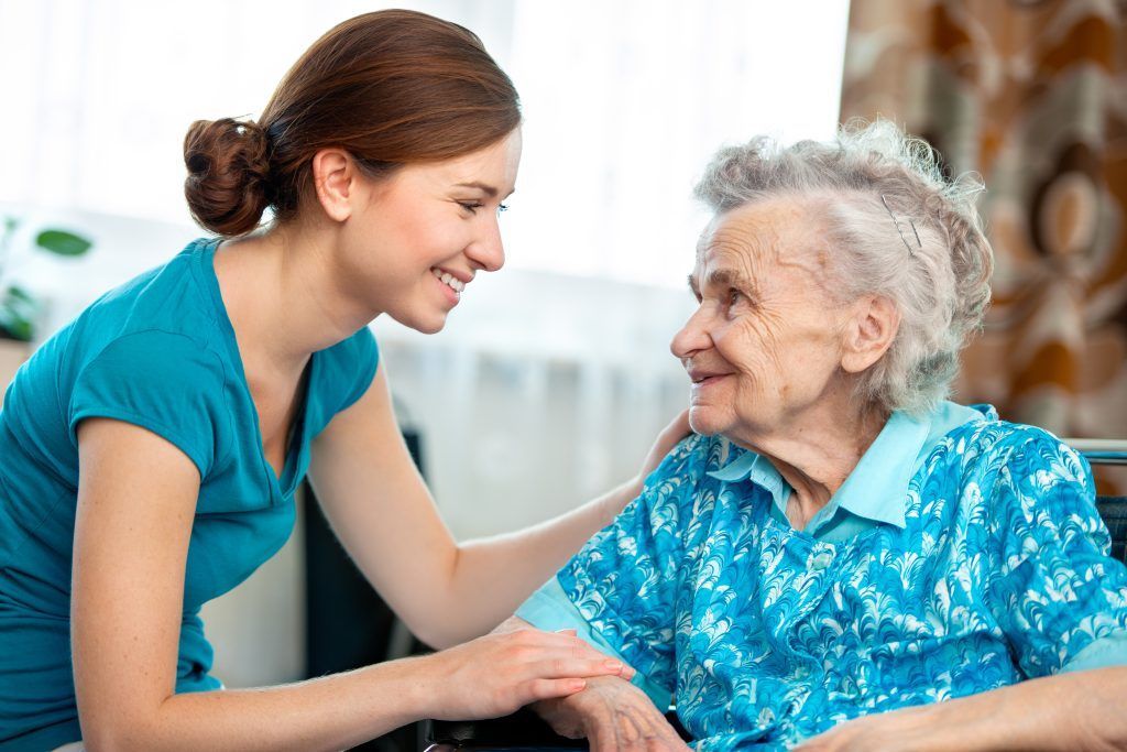 Woman in teal shirt smiles at elderly woman in blue shirt, indoors.