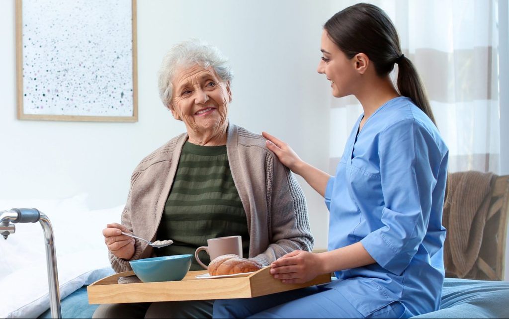 Woman in scrubs assists elderly person eating breakfast in a bedroom.