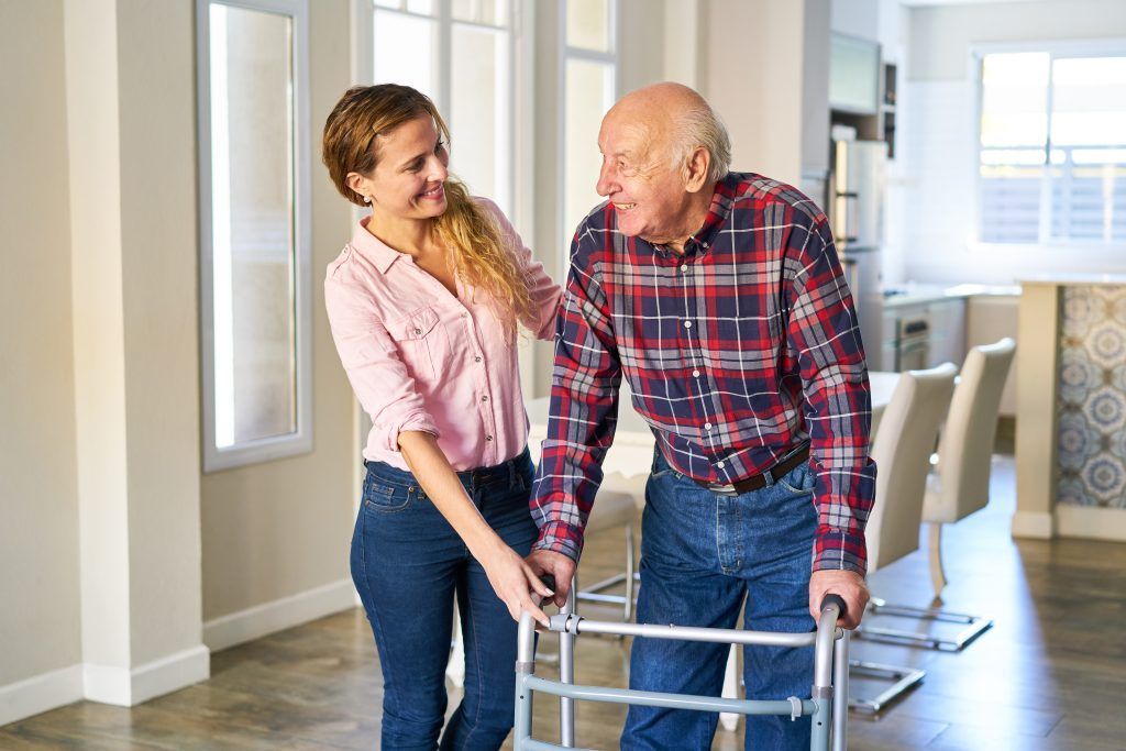 Woman assists older man using a walker in a home. They are smiling.