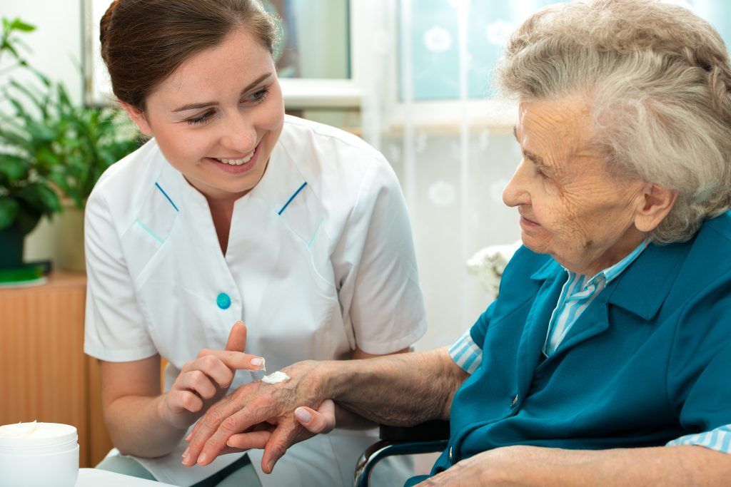 A healthcare worker applies lotion to an elderly person's hand, indoor setting, both smiling.
