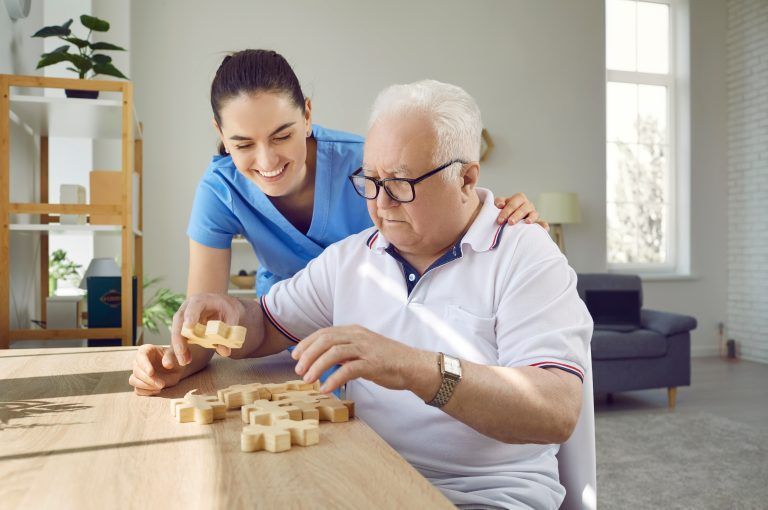 Caregiver assisting an older person with a puzzle at a table indoors.