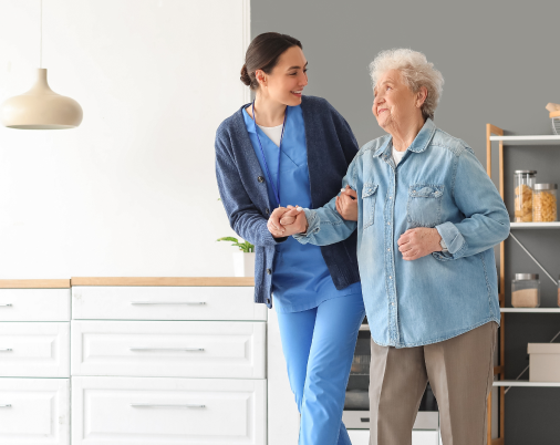 Caregiver assisting an older person walking in a kitchen, supporting their arm; smiling.