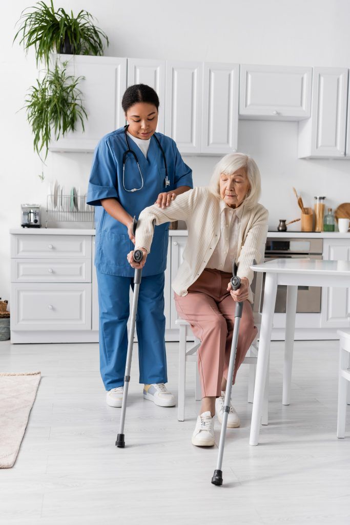 Caregiver helping a person with crutches in a kitchen. The person is sitting and the caregiver is assisting.