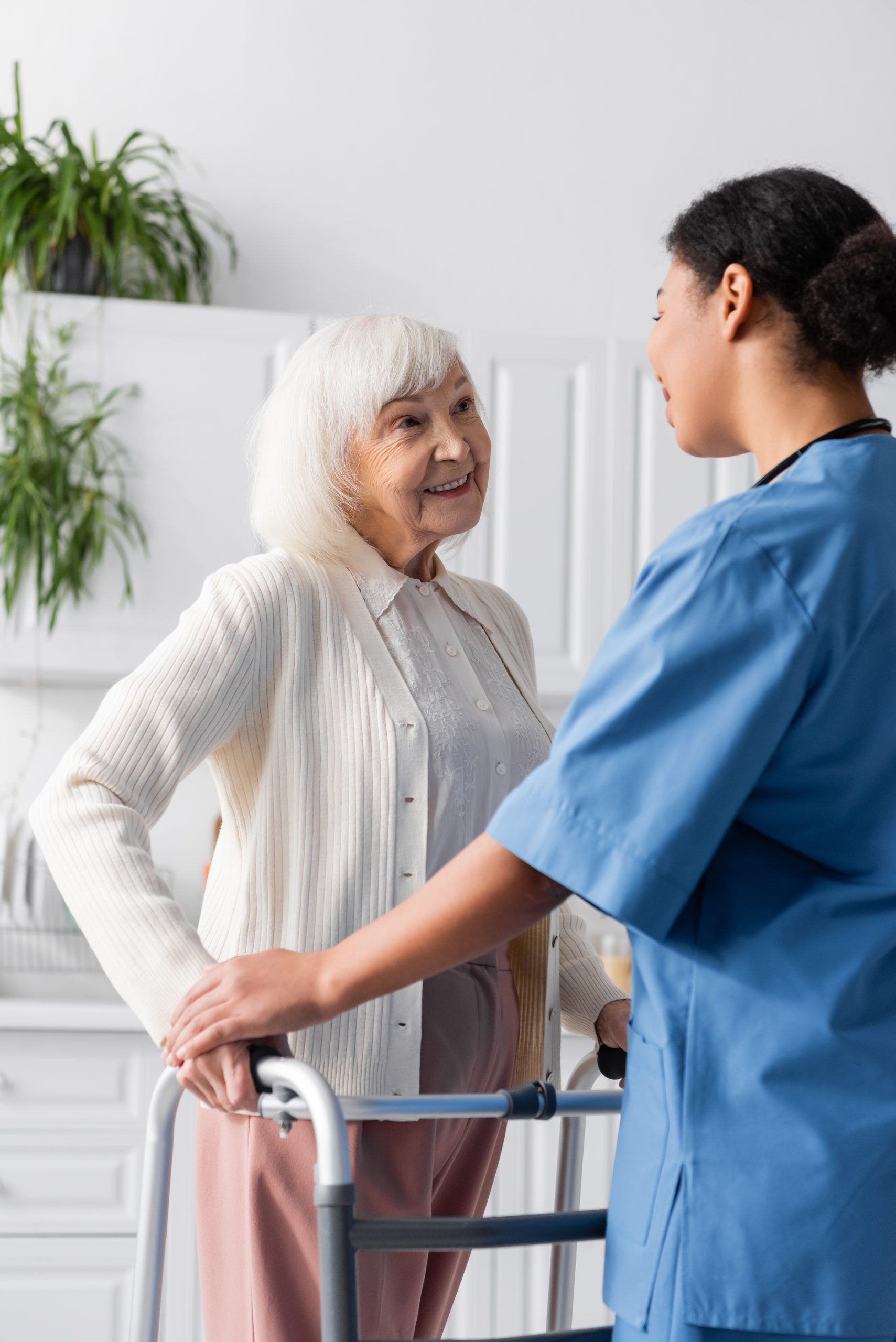 Nurse assisting a person using a walker. White sweater, blue scrubs, indoor setting.