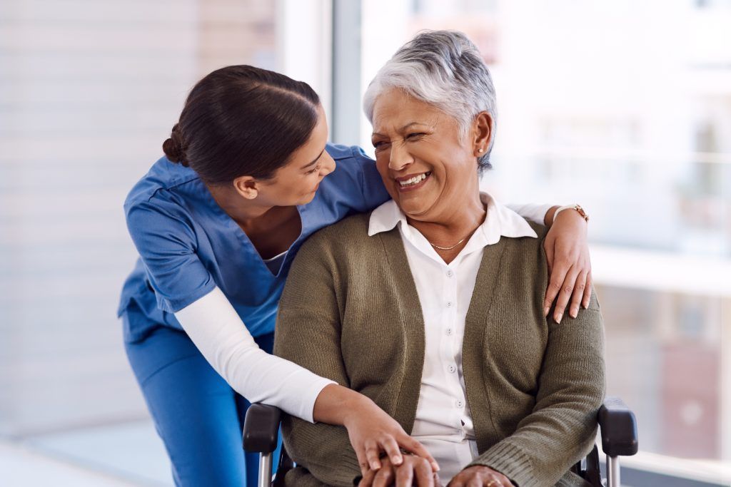 A caregiver in blue scrubs smiles at an older adult seated in a wheelchair, indoors.