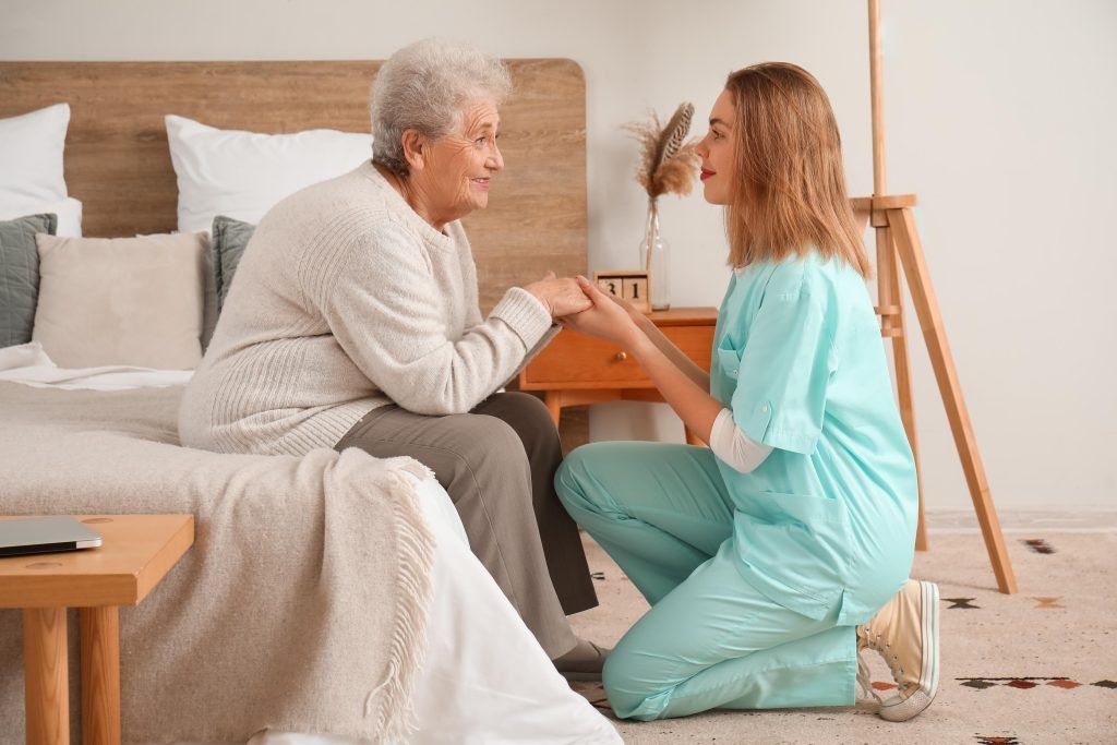 A person in scrubs kneels, holding hands with an older person sitting on a bed.