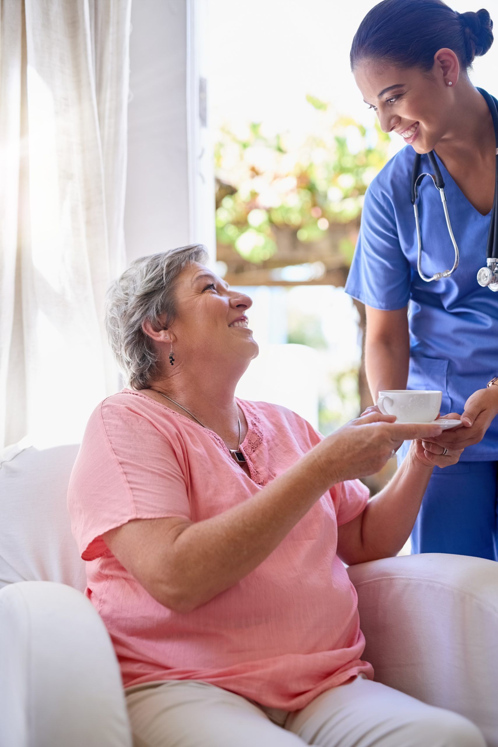 Nurse offering a cup of tea to a seated person; both smiling. Bright, interior setting.