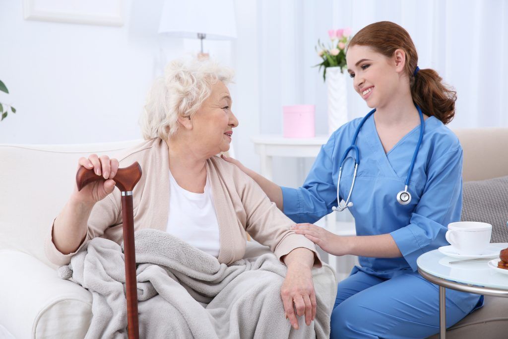 A healthcare provider with a stethoscope comforts an older person with a cane on a couch.